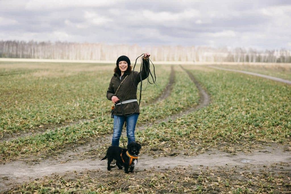 Eine Person steht auf einem Feld und hält eine Hundeleine. Ein kleiner schwarzer Hund steht neben ihr. Im Hintergrund Bäume unter bewölktem Himmel.