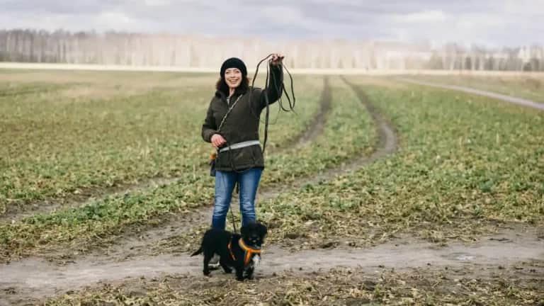 Eine Person steht mit einer Hundeleine auf einer Wiese, zu ihren Füßen liegt ein kleiner schwarzer Hund. Bedeckter Himmel.