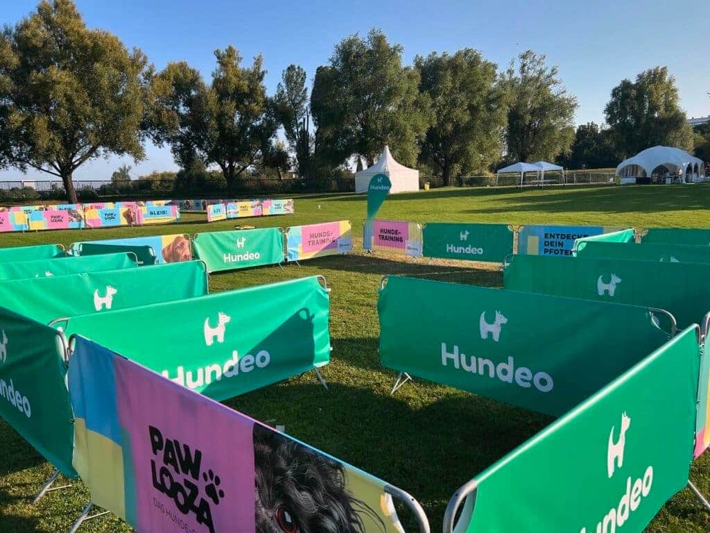 Inmitten der offenen Grasfläche erwachen eingezäunte Bereiche mit der Aufschrift „Hundeo" zum Leben und der lebhafte Geist des Pawlooza-Festivals. Bunte Banner mit Hundemotiven flattern im Wind. Große Bäume und weiße Zelte deuten auf eine große Freiluftveranstaltung hin, die dem Hundetraining und einer Feier gewidmet ist.