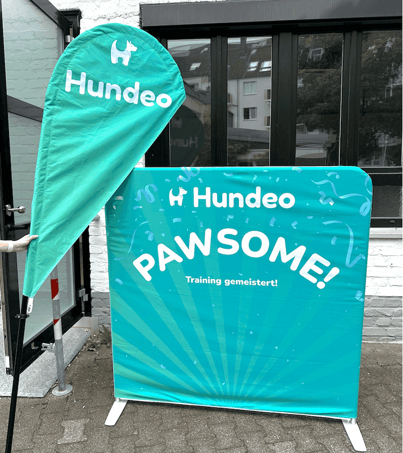 Ein blaugrünes Werbebanner und eine Flagge mit dem Text „Hundeo PAWSOME! Training gemeistert!" werden im Freien aufgestellt und fangen den festlichen Geist des Pawlooza-Festivals ein. Das Banner zeigt verspielte Pfotenabdruck-Designs vor einer Ziegelwand und einem Fensterhintergrund.